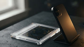 A smartphone on a small stand photographing a Pokémon card in a toploader on a plain dark background next to a window