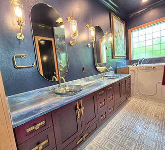 Long burgundy double vanity with brass hardware, blue-veined countertop, glass vessel sinks and oval mirrors