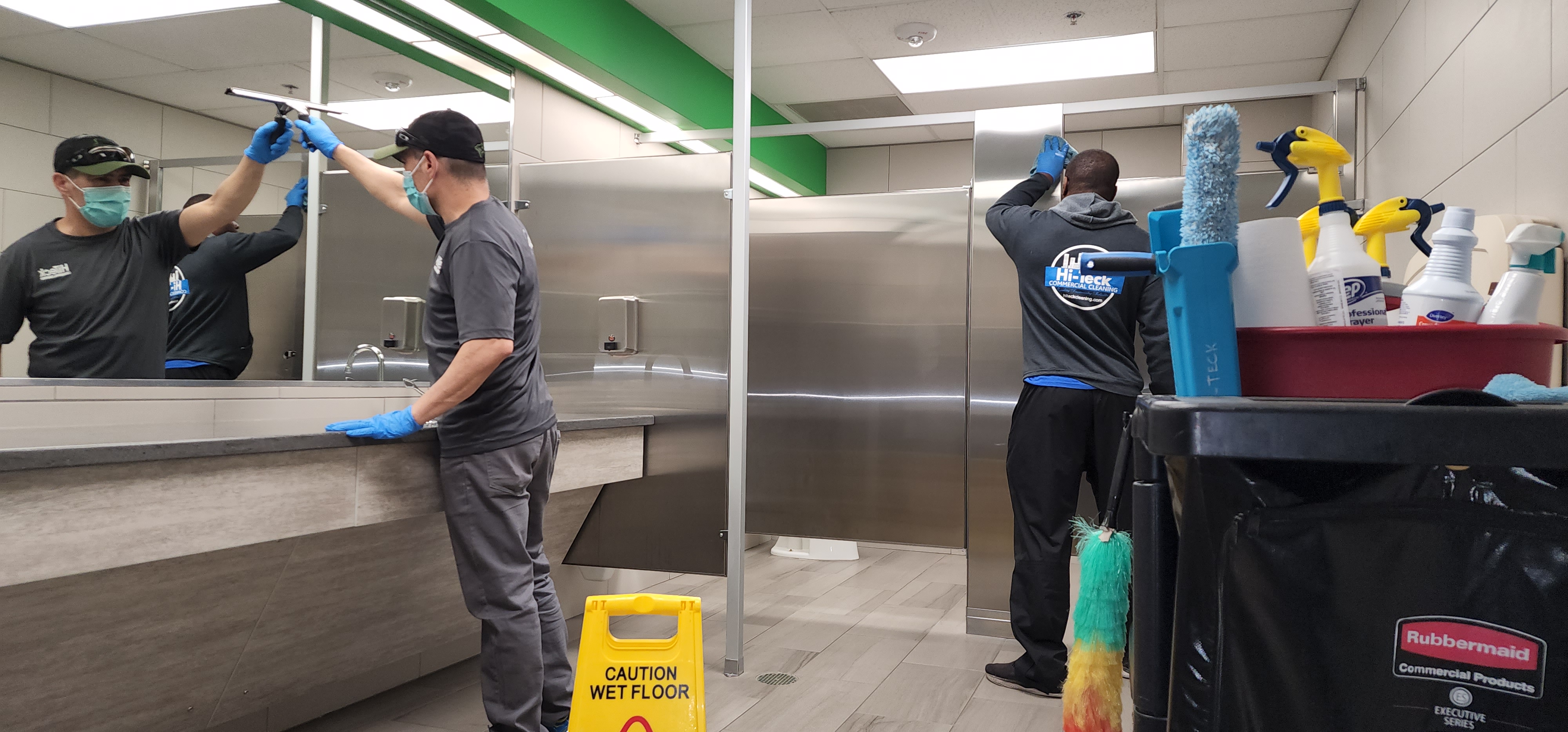 Two Hi-Teck crew members cleaning a commercial restroom, one squeegeeing a mirror and one wiping a stall divider