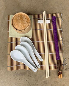 Sushi-making equipment on a bamboo mat: a bamboo steamer, white porcelain spoons, chopsticks on a rest and a rolling stick