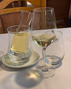 A glass carafe of white wine chilling in a glass cooler next to a wine glass and a tumbler on a white tablecloth