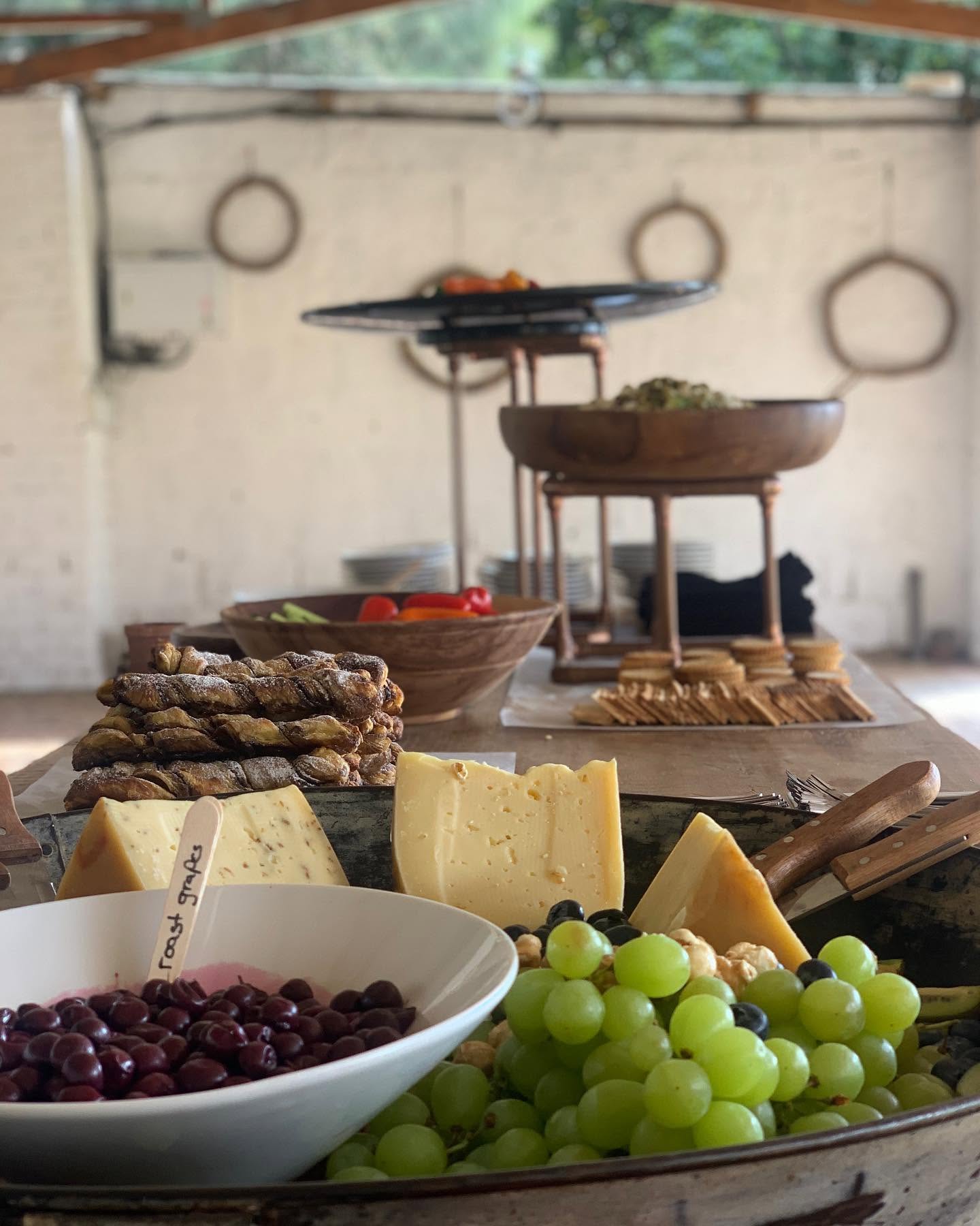 A cheese platter with wedges of cheese, green grapes, roast grapes and cheese straws, with a buffet table set up behind