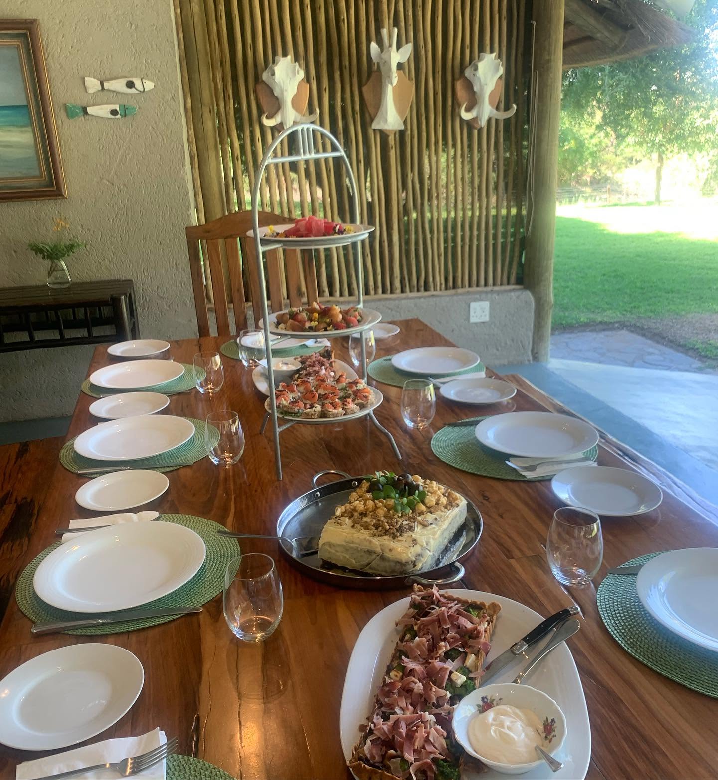 A laid timber table on a thatched stoep with white plates, green placemats, glasses, a tiered stand of canapés and a quiche