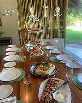 A laid timber table on a thatched stoep with white plates, green placemats, glasses, a tiered stand of canapés and a quiche