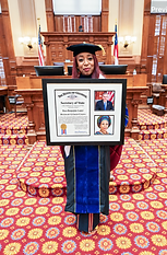 Dr. Adedayo Benjamins-Laniyi in cap and gown on the floor of the Georgia legislative chamber, holding her framed citation naming her an Honorary Georgia Citizen, signed by Secretary of State Brad Raffensperger