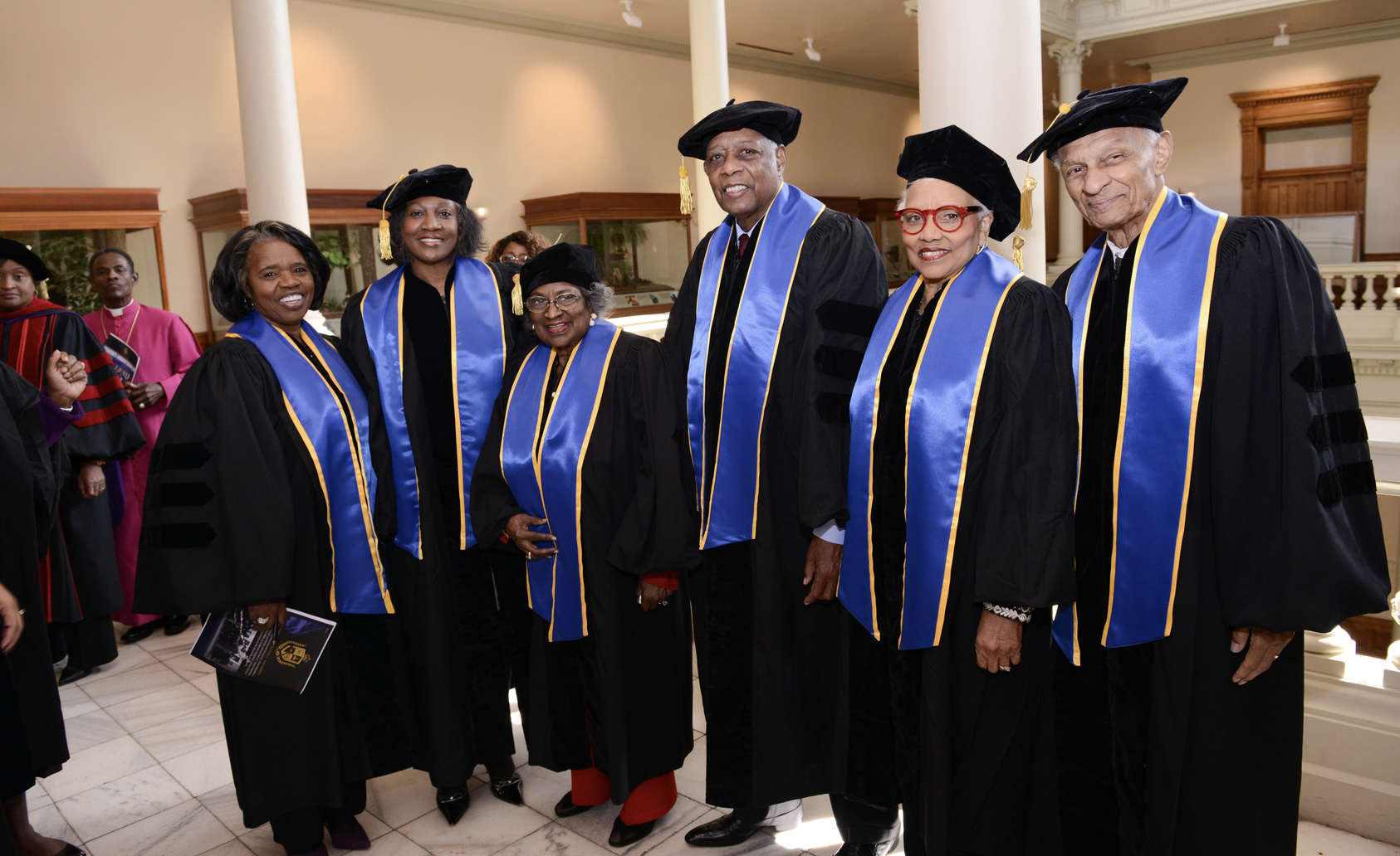 Six TIUAC graduates posed together in caps, gowns, and blue stoles