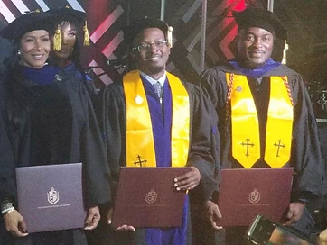 Three TIUAC doctoral graduates in caps and gowns on stage holding maroon Trinity International University of Ambassadors diploma covers