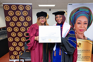 Dr. Adedayo Benjamins-Laniyi and a colleague in caps and gowns holding her framed Doctor of Philosophy certificate before a congratulatory banner