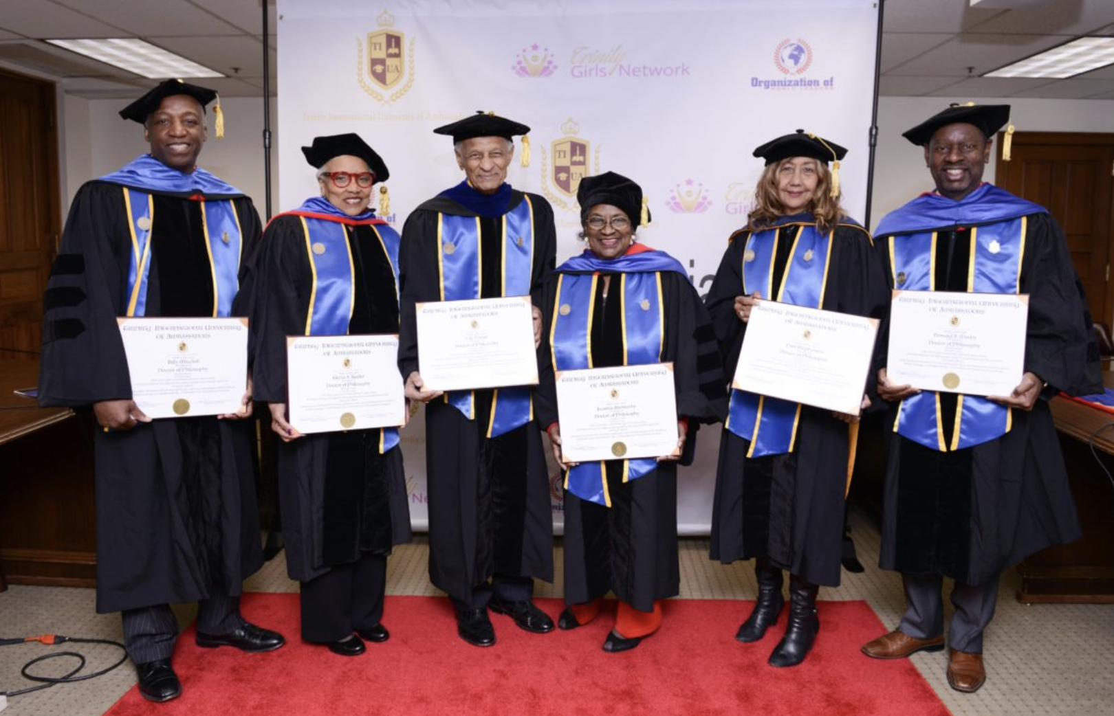 Six TIUAC graduates holding framed diplomas before the Trinity International University of Ambassadors backdrop