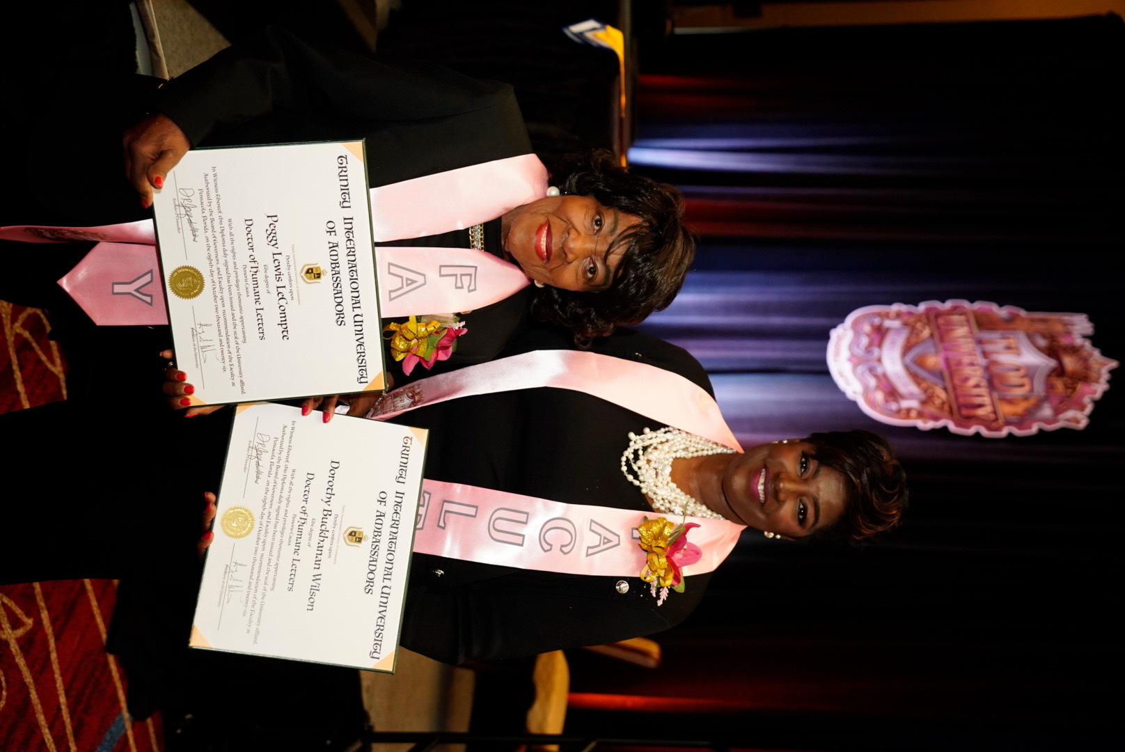 Two TIUAC honorees in formal attire with pink sashes holding their Doctor of Humane Letters diplomas