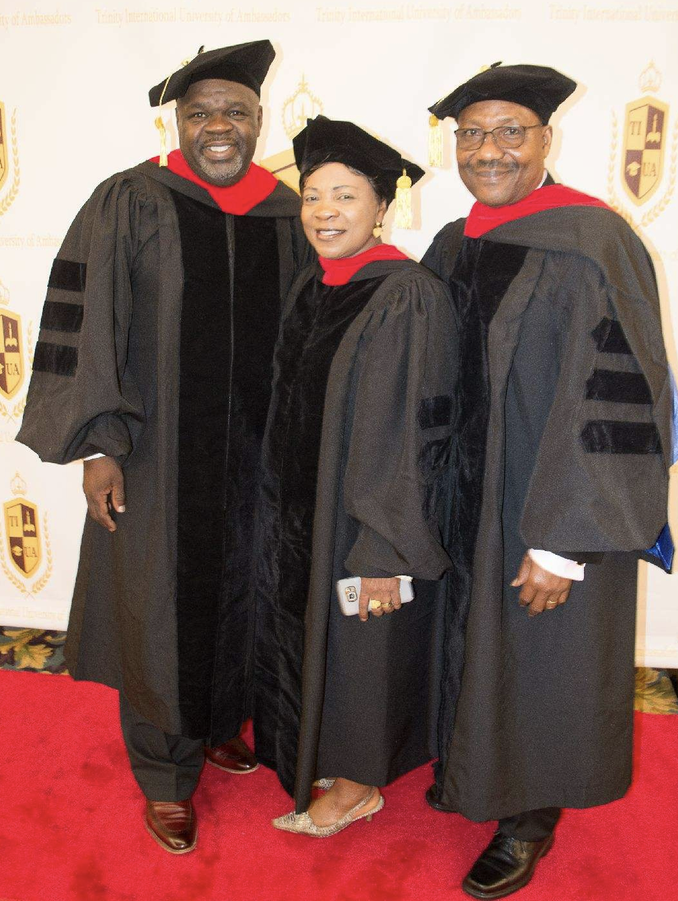 Three TIUAC doctoral graduates on the red carpet before the university crest backdrop