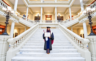 Dr. Adedayo Benjamins-Laniyi in academic regalia standing at the center of a grand marble staircase, holding a framed certificate and an American flag
