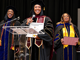 A graduate in a black tam and doctoral gown with blue velvet panels speaking at the ceremony lectern, flanked by two graduates in academic regalia