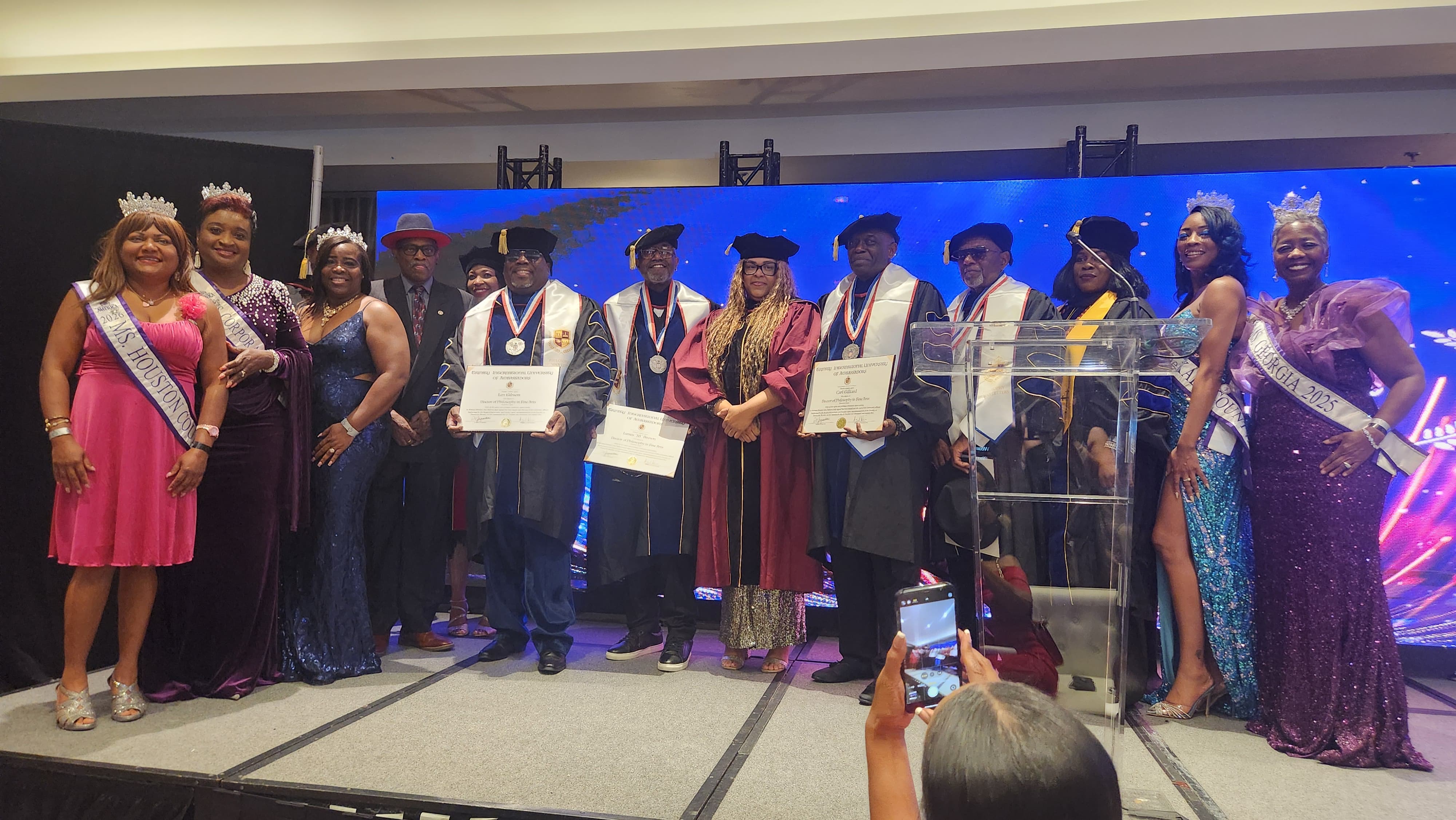 Honorees in academic regalia and pageant sashes holding Trinity International University of Ambassadors certificates on stage
