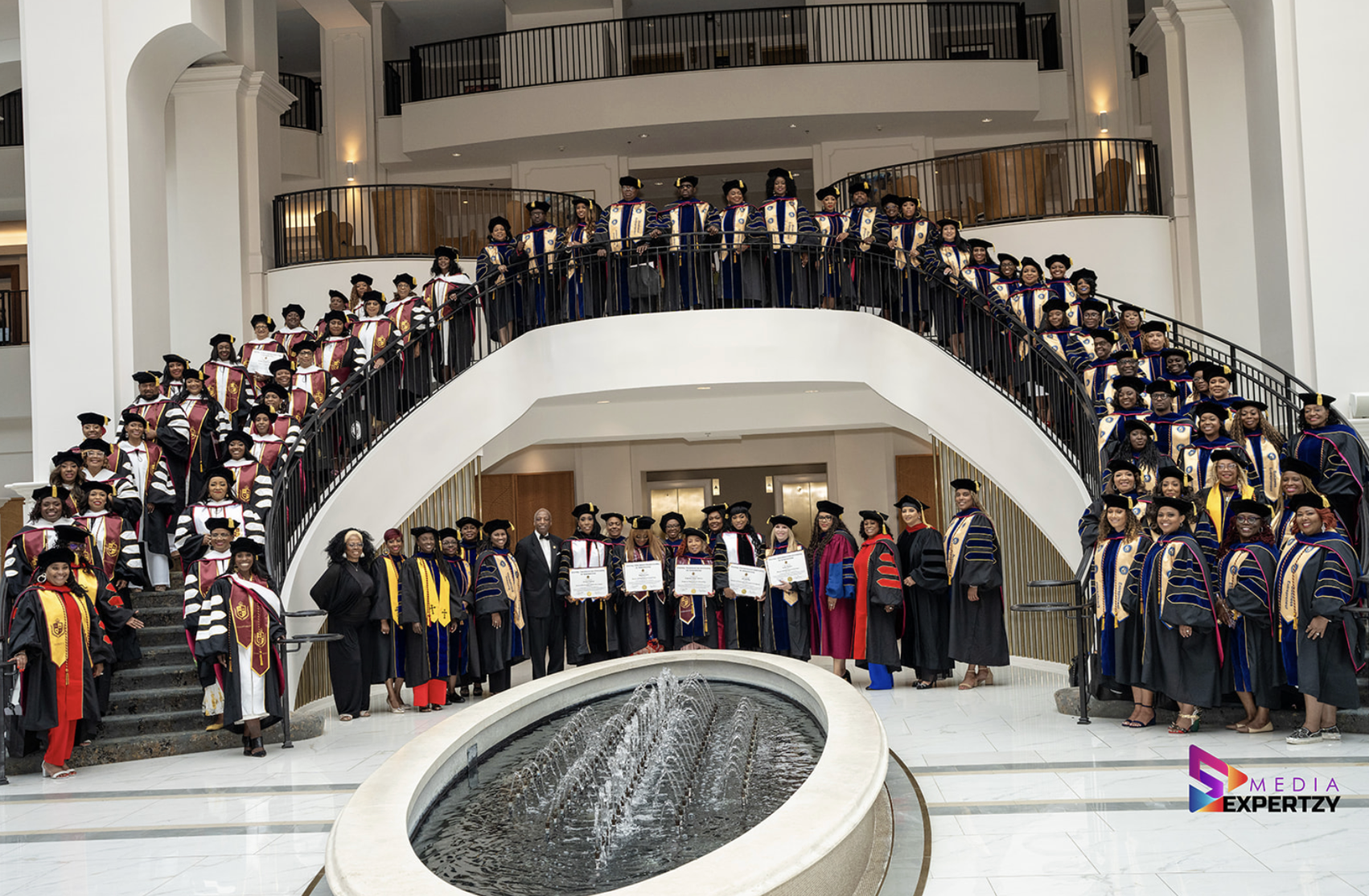 Large assembly of TIUAC graduates in caps and gowns on a grand staircase beside a fountain