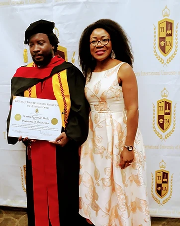 A TIUAC doctoral graduate in regalia holding his Doctorate of Philosophy diploma with a guest before the Trinity International University of Ambassadors backdrop