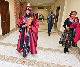 Dr. Adedayo Benjamins-Laniyi in maroon academic regalia carrying a bouquet of flowers and a small American flag down a capitol hallway