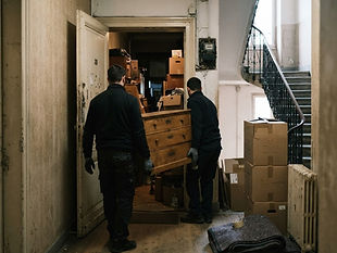 Entrümpelung Wohnung: Wohnungsräumung: zwei Mitarbeiter tragen eine Kommode aus der Wohnung, Kartons im Flur.