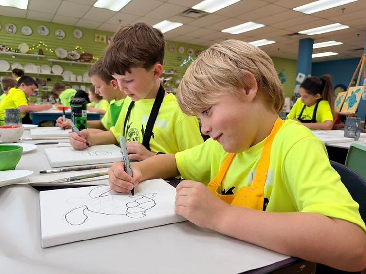 Kids in neon camp shirts and aprons drawing on canvases during a creative camp activity