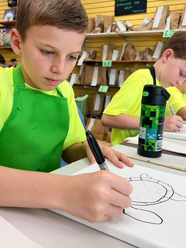 A camper in a green apron focused on drawing a sea turtle on canvas