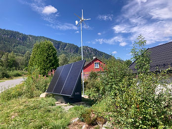 SoWin Kraftstasjon med solcellepanel og vindturbin i eng ved rød hytte og skogkledd fjell