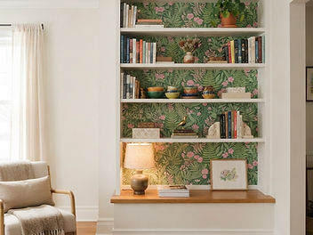 White built-in bookcase with the back of each shelf lined in green and pink botanical wallpaper, styled with books, ceramics, a table lamp and a small plant beside a cream armchair