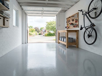 Bright single garage with a glossy light grey epoxy floor, white painted brick walls, an oak workbench with tools on a pegboard and a bicycle hung on the wall, with the up-and-over door open to daylight