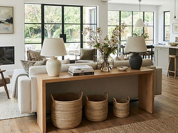 Long slim oak sofa table behind a floating linen sofa in a bright open plan living room, with two ceramic table lamps, books, a vase of branches and woven baskets underneath