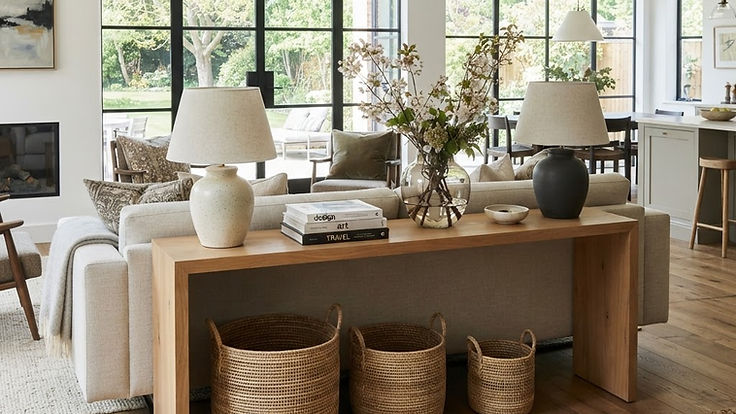 Long slim oak sofa table behind a floating linen sofa in a bright open plan living room, with two ceramic table lamps, books, a vase of branches and woven baskets underneath
