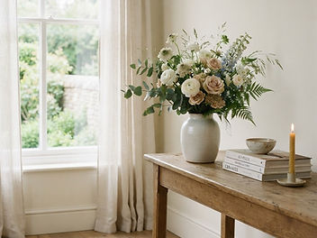 Fresh bouquet of white and blush roses with blue flowers in a ceramic vase on a rustic wooden console table by a sunny window