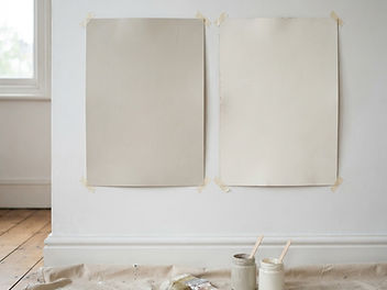 Two large painted sample boards side by side on a white wall, one a slightly darker warm greige off-white and one a brighter creamy white, with a paintbrush and small paint pots on a dust sheet below