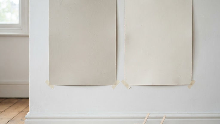 Two large painted sample boards side by side on a white wall, one a slightly darker warm greige off-white and one a brighter creamy white, with a paintbrush and small paint pots on a dust sheet below