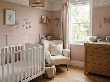 Soft baby girl nursery with dusty blush pink walls and panelling, a white cot with a clear mattress, a cream boucle nursing chair, an oak chest of drawers used as a changing station and a sash window with floral curtains