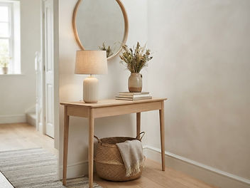 Scandinavian hallway with a slim pale oak console table on tapered legs, a round oak mirror, a ribbed ceramic lamp with a linen shade, a vase of dried grasses, a stack of books and a woven basket underneath