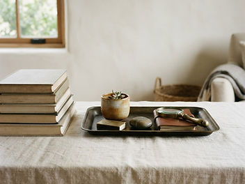 Neatly styled linen-covered table with a stack of books and a dark tray holding a small plant and everyday essentials