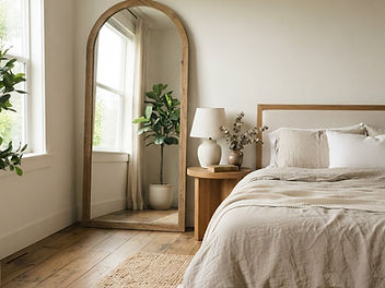 Serene bedroom with a huge arched floor mirror leaning against the wall beside the bed, reflecting window light, with linen bedding, an oak bedside table and warm white walls