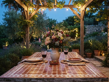 Rustic outdoor dining table with a terracotta and white zellige tiled top under a pergola with string lights, set with plates and a jug of garden flowers in warm summer evening light