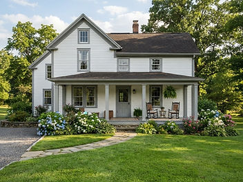 Classic white farmhouse with white clapboard siding, mid-gray painted window trim and porch posts, a wide front porch with rocking chairs, a gray front door, hydrangeas and a green lawn
