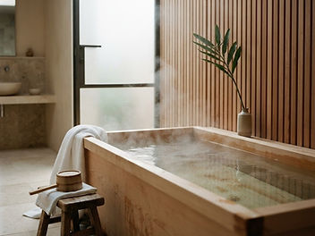 Steaming hinoki wood Japanese soaking tub beside a timber bath stool with a wooden bucket and white towel, in a calm bathroom with slatted wood walls and a frosted glass door