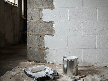 Basement concrete block wall half painted with white masonry waterproofing paint, with a paint roller in a tray, an open paint tin and dust sheets on the floor