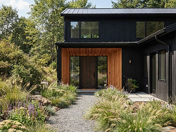 Modern black house with vertical black siding, black window frames and a warm cedar-clad entrance with a wooden front door, reached by a gravel path through ornamental grasses