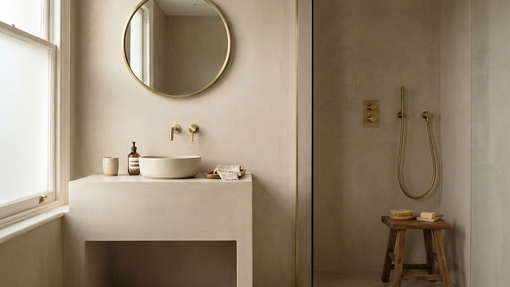 Spa-like bathroom finished in seamless warm greige microcement on the walls, floor and a built-in vanity, with a walk-in shower, brushed brass fittings, a round mirror and a wooden stool in soft natural light