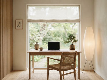 Japandi home office with a light oak desk facing a garden window with a linen blind, a woven rattan chair, a paper floor lamp, a slatted oak wall panel, a bonsai and a vase of blossom