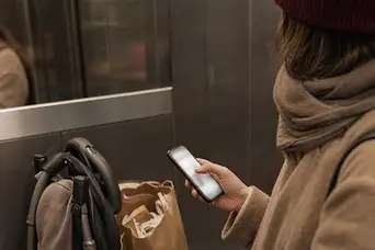 Femeie în liftul blocului, cu telefonul în mână, o sacoșă și căruciorul pliat alături.