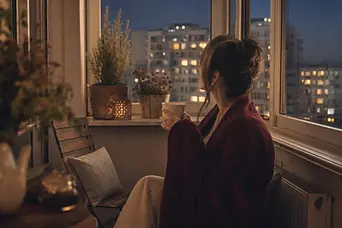 Femeie pe balconul închis, seara, cu o cană în mâini, privind ferestrele blocului de vizavi.