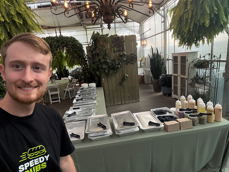 Aidan at a catered event setup with pizzas, subs, and sides