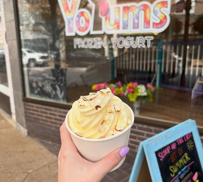 A hand holding a paper cup of frozen yogurt topped with sliced almonds and shredded coconut, in front of the YoYums storefront on East High Street.