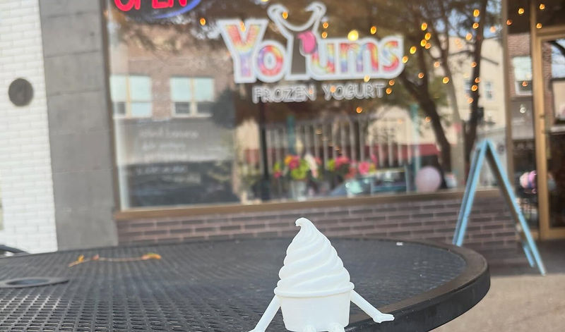 A small white figurine shaped like a soft-serve swirl sitting on a patio table outside the YoYums storefront, the shop windows glowing softly behind it.