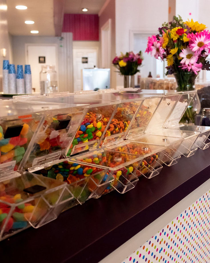 The toppings bar at YoYums: clear angled bins packed with gummy worms, chocolates and jelly beans, glass jars of sprinkles behind them, and a bouquet of pink and yellow daisies on the counter.