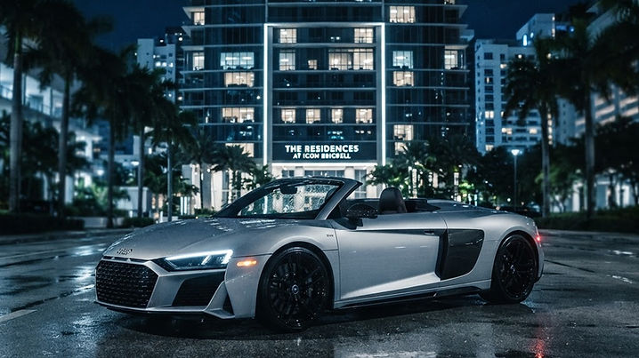 A silver Audi R8 Spyder convertible parked at night in front of a sleek Miami residential high-rise